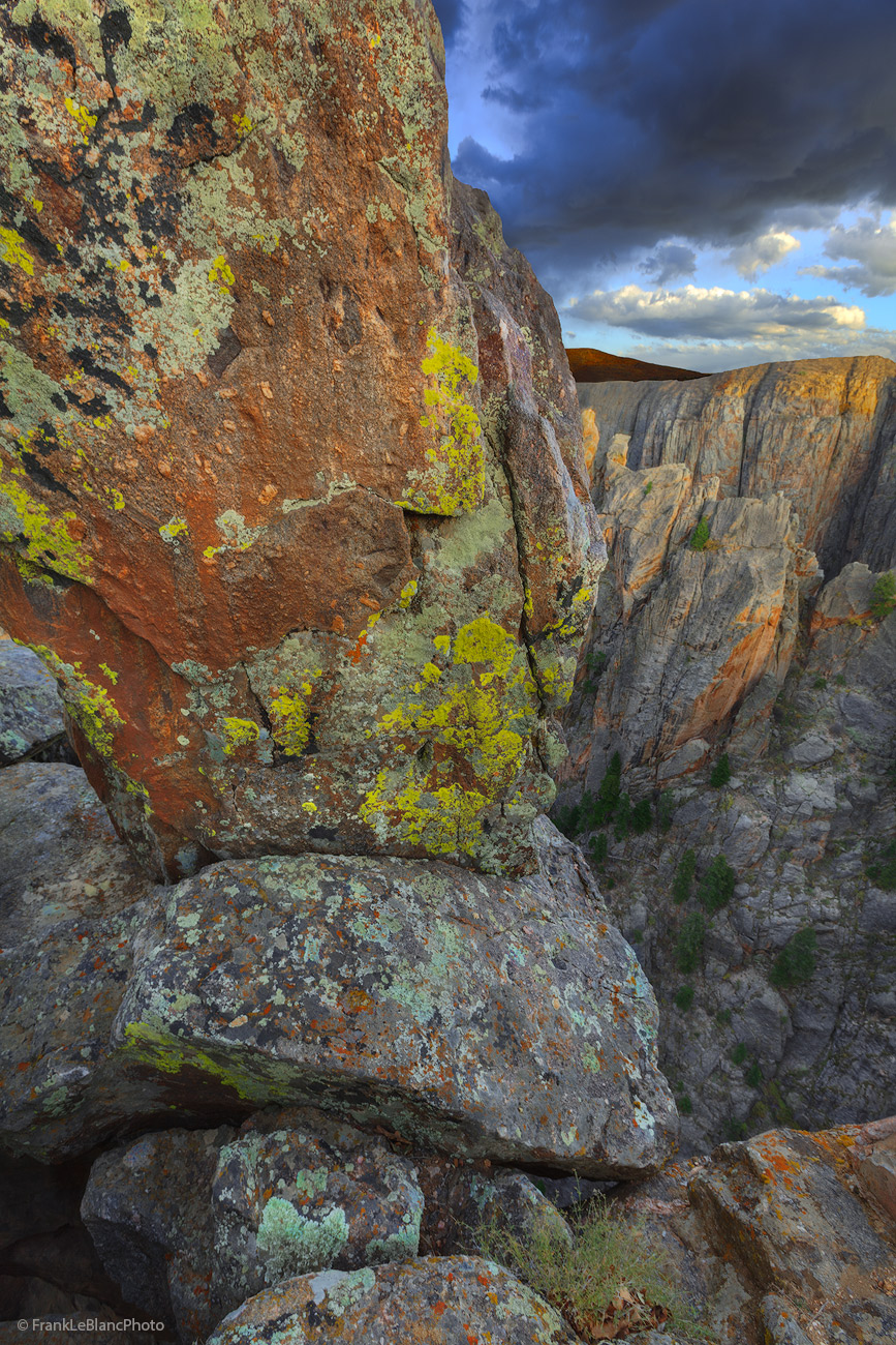 Storm clouds sweep over the abyss of Black Canyon at sunset. Huge rocks brightly painted with lichens and fungi totter on the...