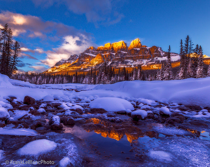 Castle mountain is reflected in a small pool of water surrounded by hoarfrost and ice. The Bow river is hidden by the mid-ground...
