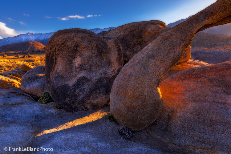Setting sun is casting intense light upon Mobius Arch, surrounding boulders, and the mountain landscape beyond.