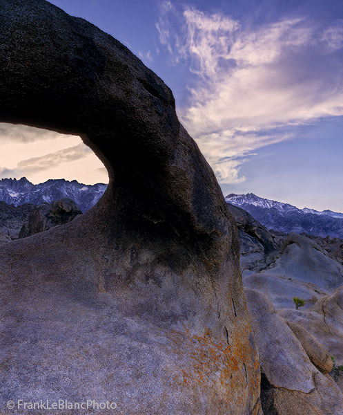 A dark and backlighted arch gives a sunset view deep into the Alabama Hills and mountains of the Sierra Nevada. Clouds move from...