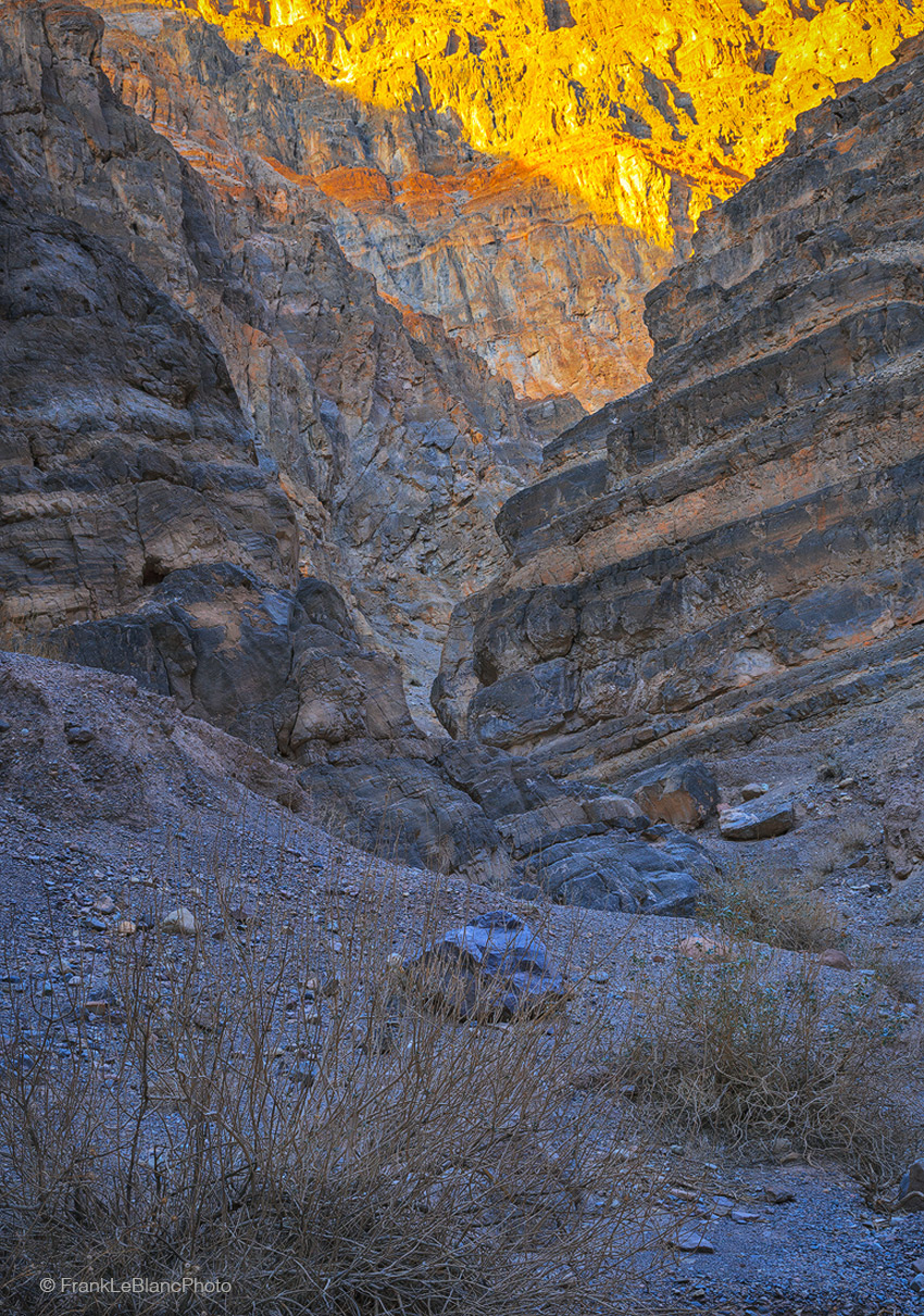 Different levels of light filter deep into Titus canyon at sunset from intense yellow orange above to dark olive brown on the...